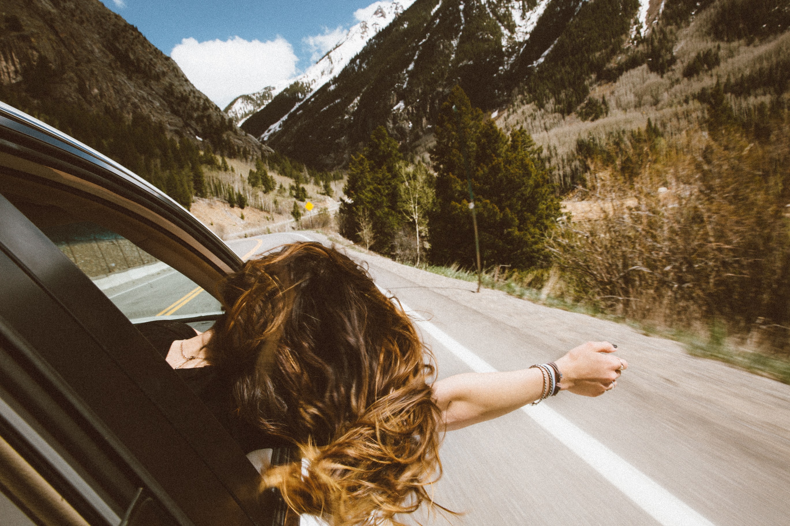 A woman hangs her head out of the car window in joy while driving along a mountain road
