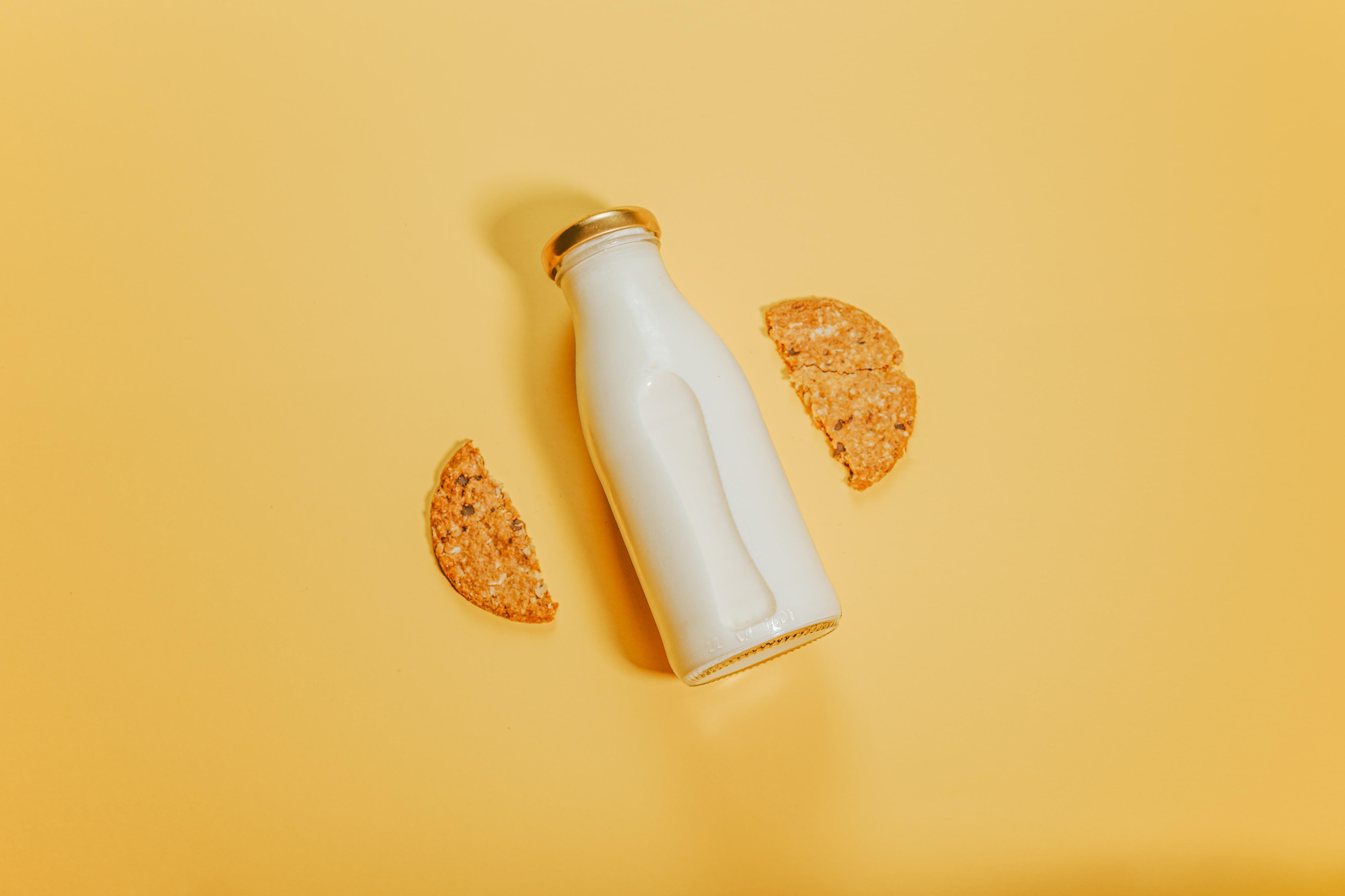 A small, glass bottle of milk against a yellow background with a cookie snapped in half