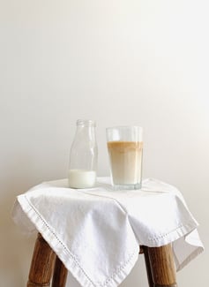 A jug of milk sits on a wooden stool with a white cloth, next to a glass of milk iced coffee