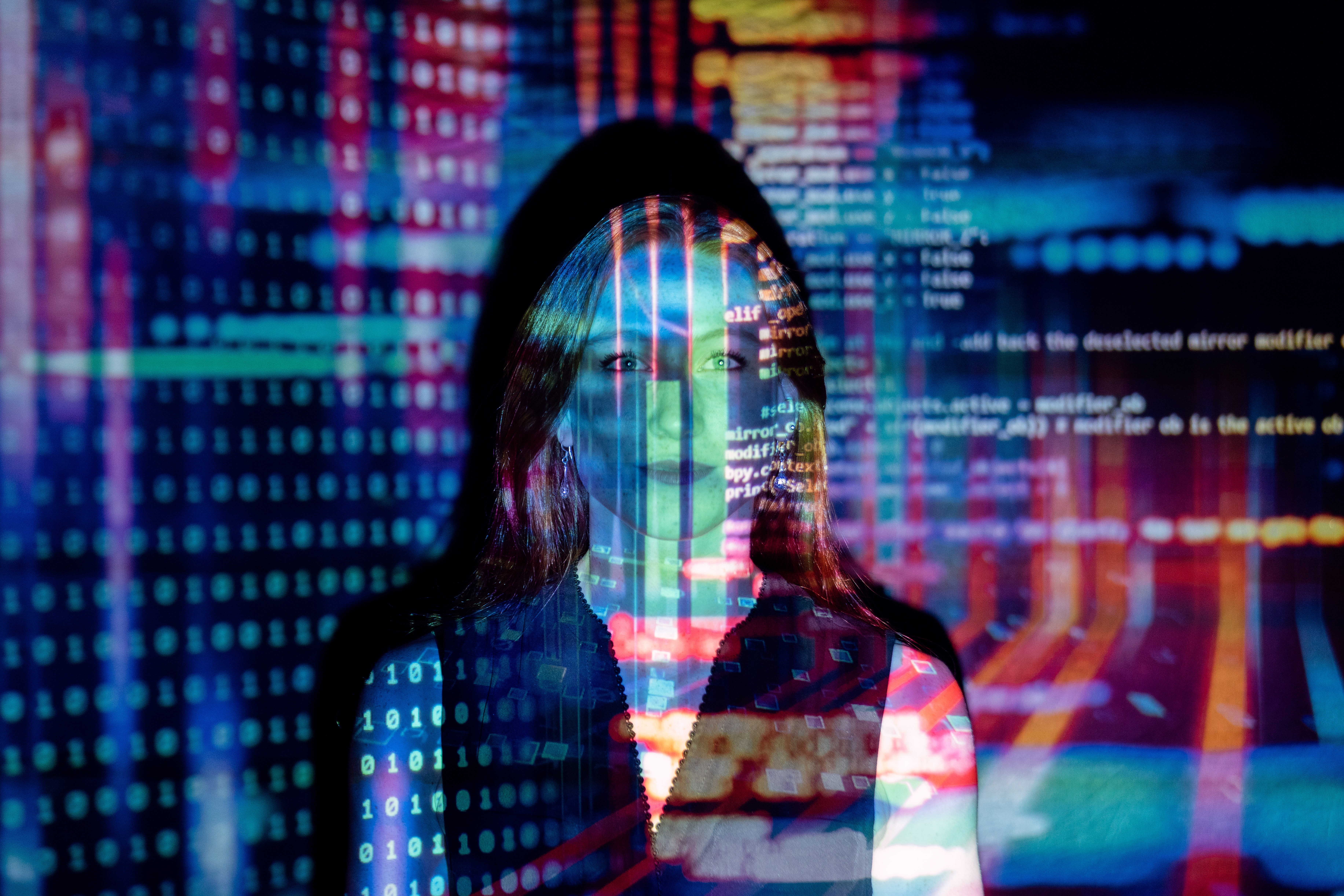 A woman stands against a wall with lines of code in multi color projected onto her