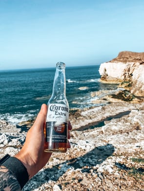 A person holds a bottle of Corona beer up against a beach background