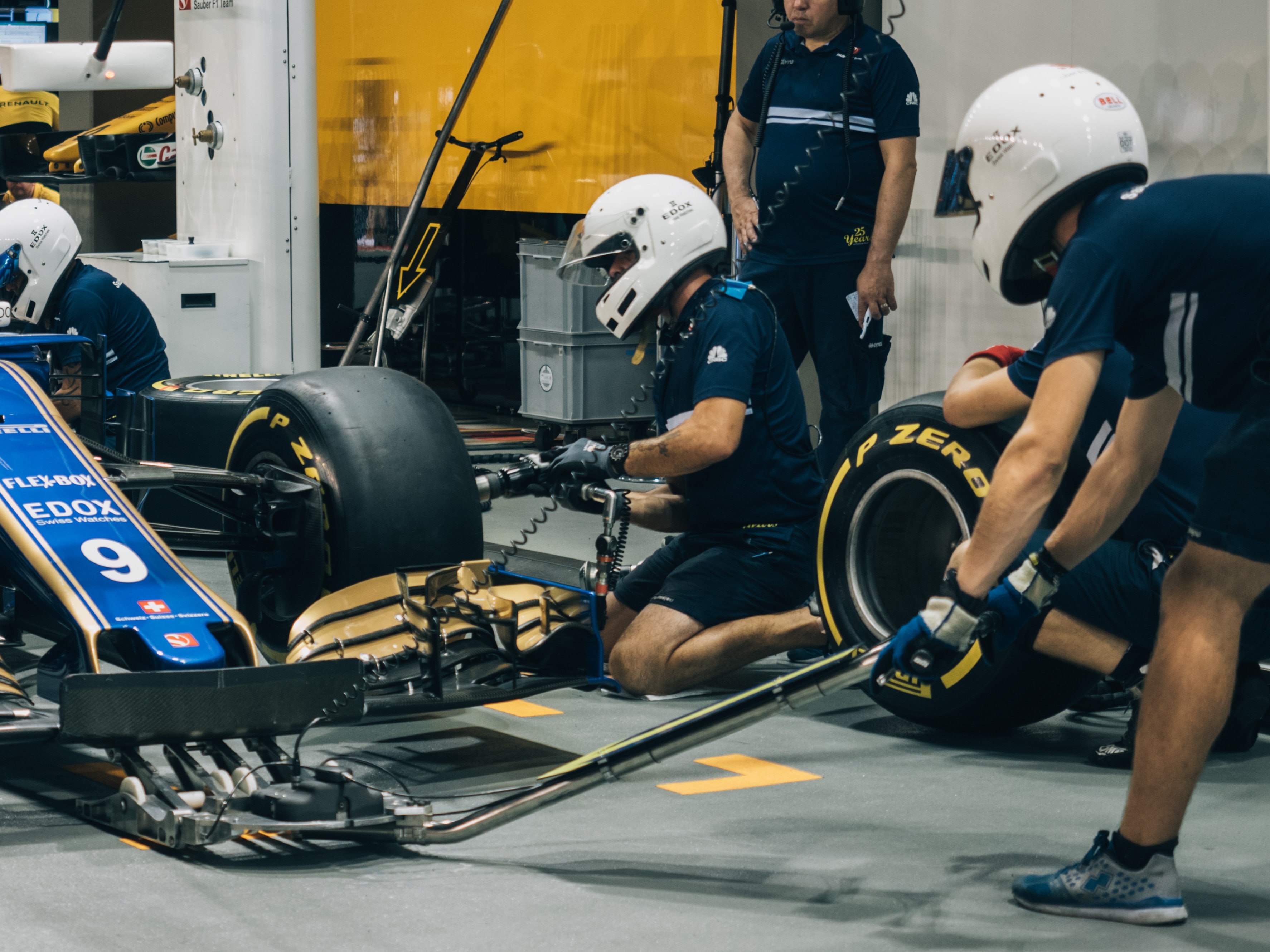 A racecar being worked on by a pit crew with white helmets. One crew member is changing a tire and the other is using a jack to lift the car up.