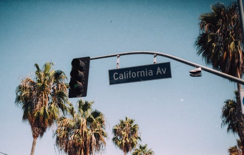 A street sign reading 'California Ave' with a soft blue sky and palm trees in the background