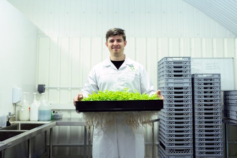 A man in a white lab coat stands smiling in a workroom, holding a tray of green plants
