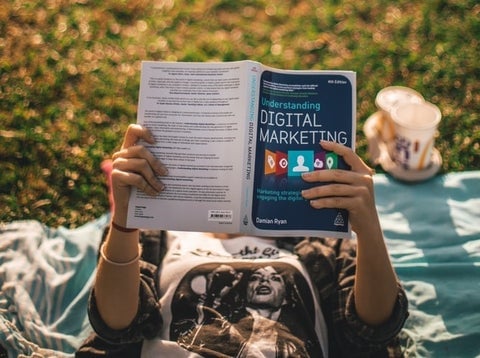 A woman lies back on a picnic blanket in the sun reading a book titled 'Digital marketing'