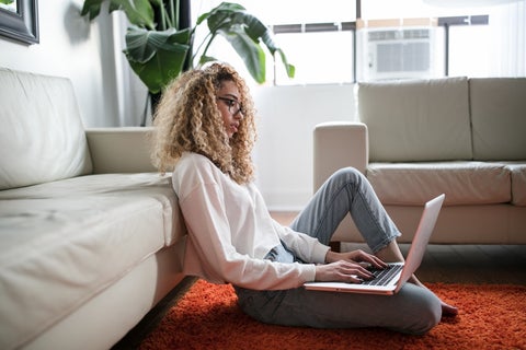 A curly haired woman sits on the floor with her laptop