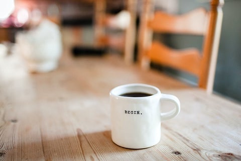 A white coffee mug on a wooden kitchen table, marked with the word 'Begin'