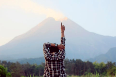 A man stands facing a rural landscape with one finger pointed up into the air