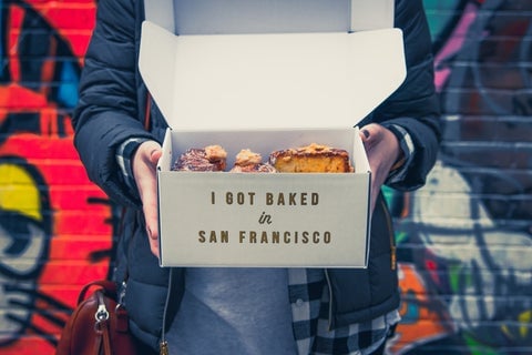 A person stands against a graffitied wall holding a box of baked goods. The packaging reads: "I got baked in San Francisco"