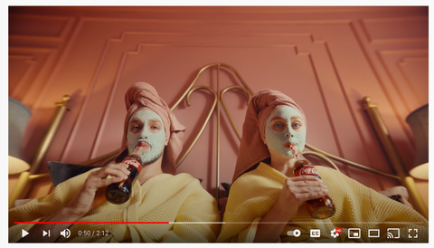 A man and a women in yellow bathrobes recline with face packs, sipping Coca-Cola through a straw