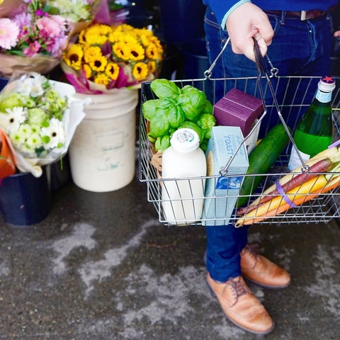 A man stands outside a food store, with a basket containing basil, milk, water, and other groceries