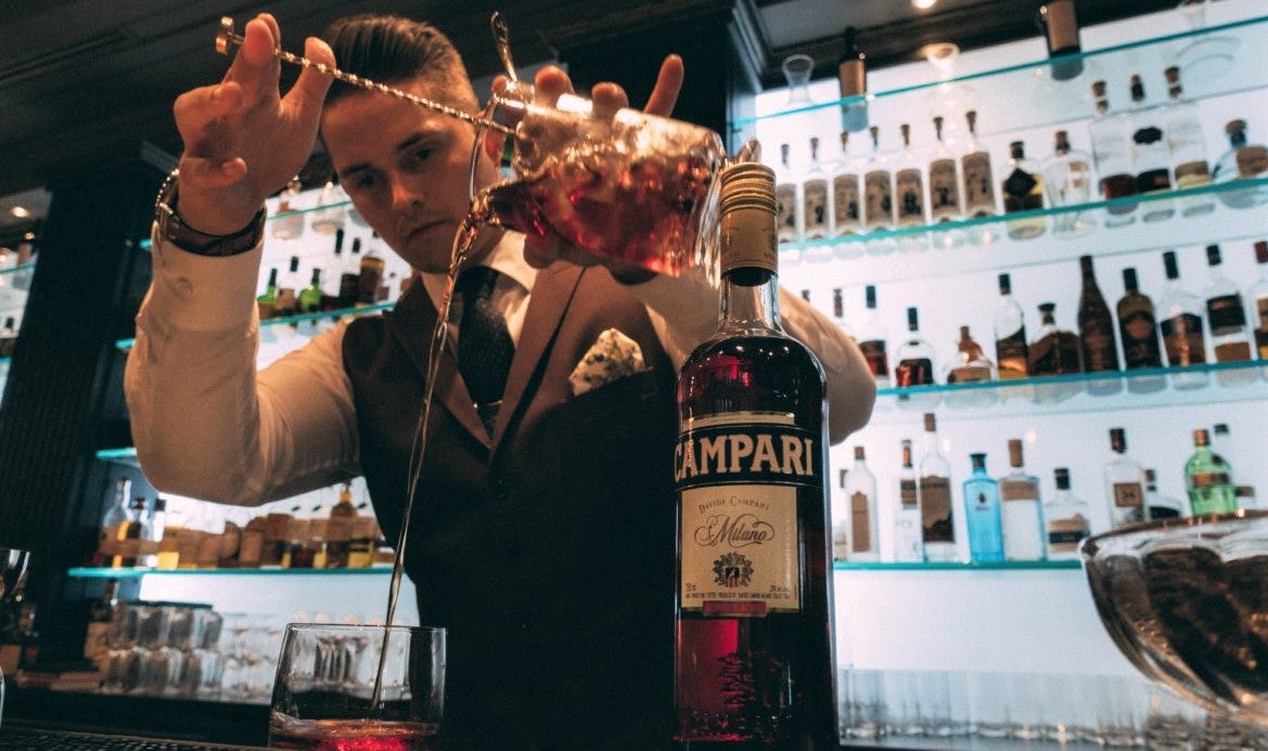 male bartender in white shirt and bowtie making cocktail against well-lit bar