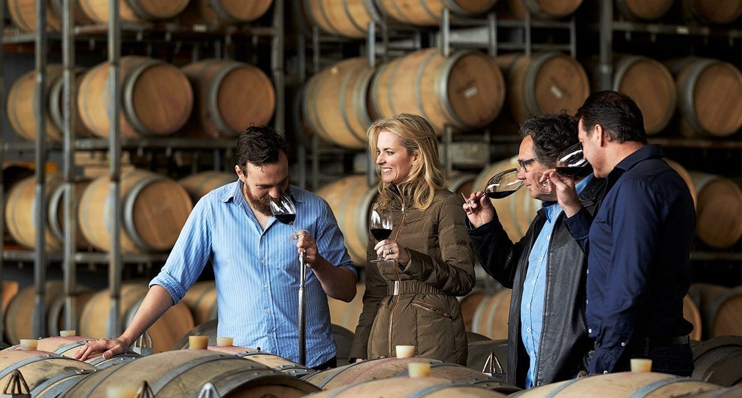group of wine makers stand at large barrel and test out new wine