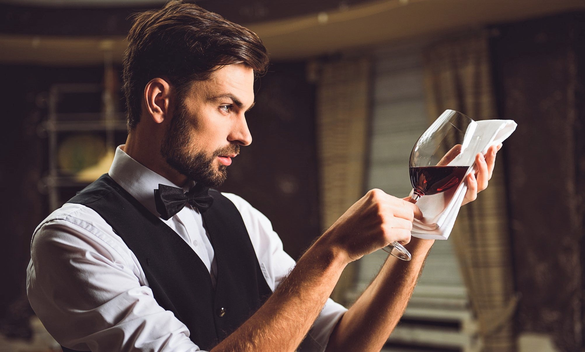sommelier in white shirt and black waistcoat inspects glass of red wine