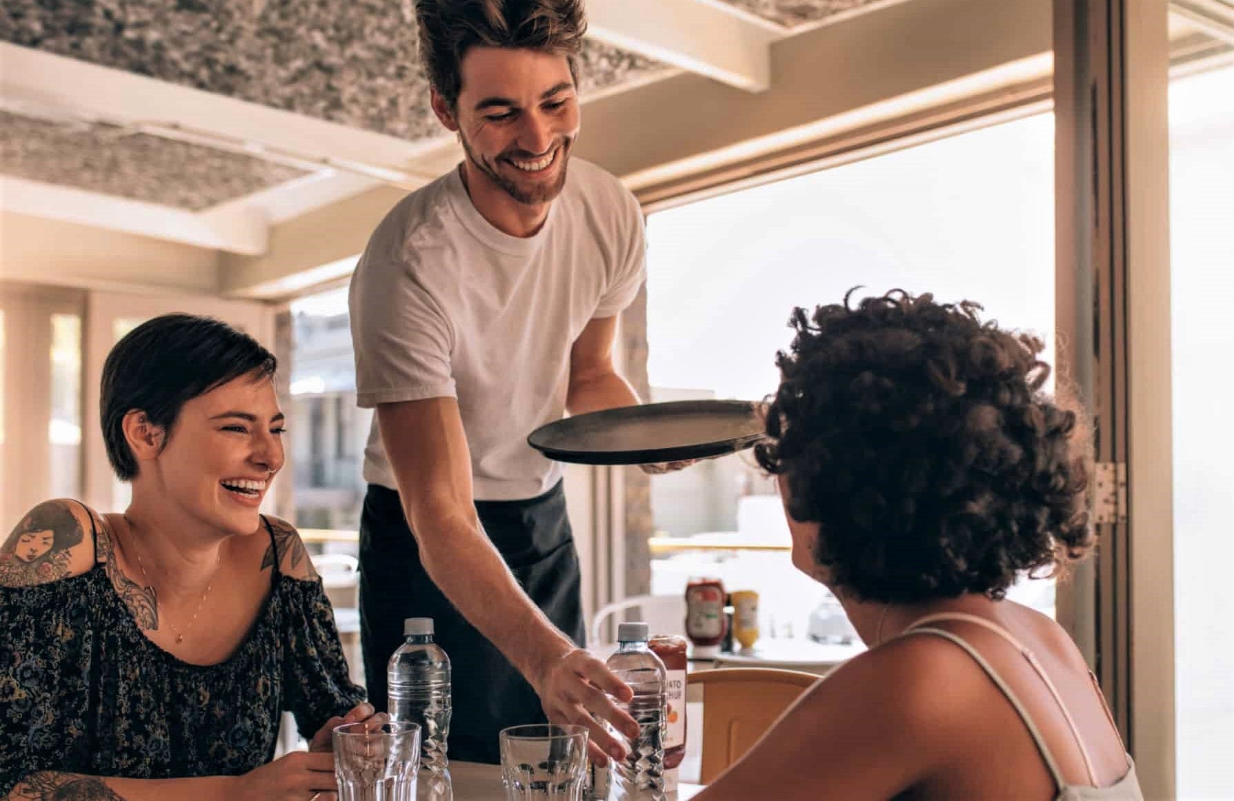 attractive young waiter in t-shirt serves two women drink at restaurant table