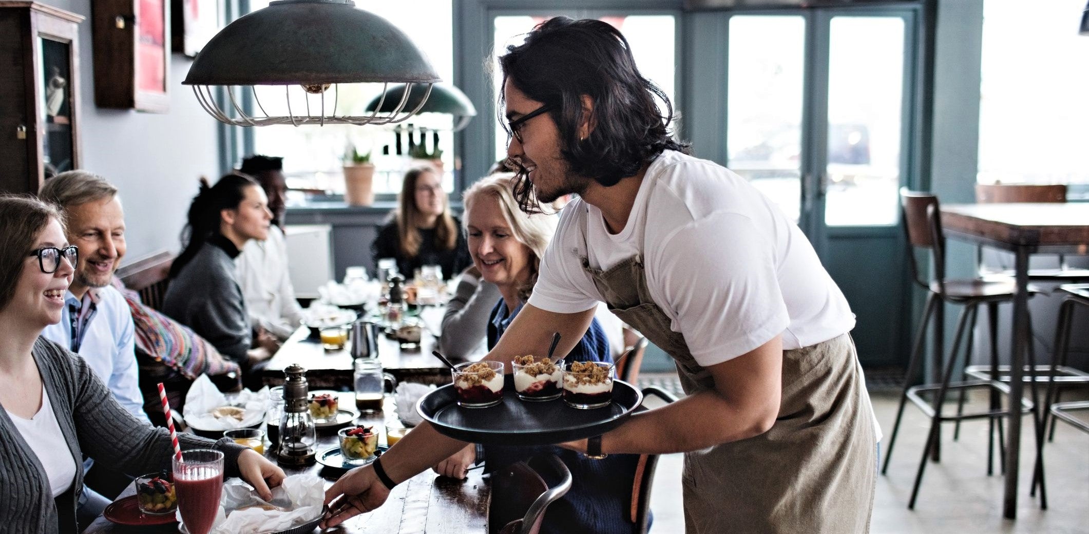 attractive young waiter with long hair and glasses delivers tray of desserts to table of smiling repeat customers